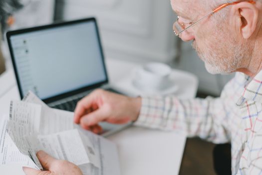 Man in eyeglasses reading a stack of documents next to a laptop at home.